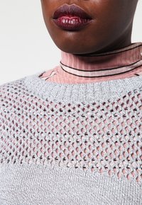Grauer gestrickter Pullover mit einem strukturierten Mesh-Design am Halsauschnitt, der über einem hellrosa gerippten Rollkragenpullover mit schwarzen Streifen getragen wird.