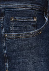 Jeans de mezclilla oscura con un solo bolsillo trasero, costuras en contraste y un pequeño detalle de remache cerca del bolsillo. Textura suave en general.