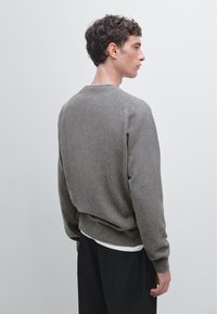 Pull gris en tricot avec un col rond, des poignets et un ourlet côtelés, vu de l'arrière, associé à une chemise blanche en dessous.