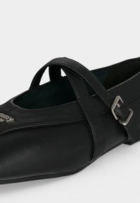 Chaussure plate en cuir noir avec un bout pointu, double brides croisées et une boucle en métal argenté sur le côté.