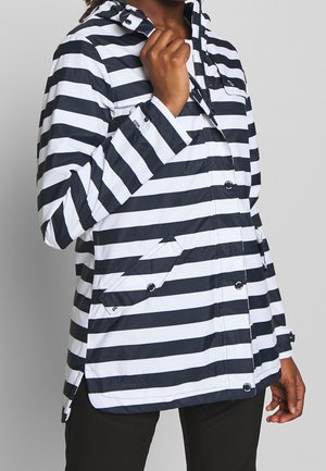 Person, die den Kragen einer schwarz-weiß horizontal gestreiften Regenjacke mit Druckknöpfen und schrägen Fronttaschen anpasst.