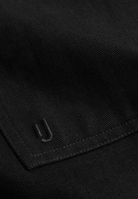 Fekete anyag, bordázott textúrával, a bal alsó sarokban egy kis hímzett logó található, azonos fekete cérnával, és egy letisztult varrással.