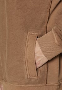 Detalle de sudadera marrón con un bolsillo lateral, puños acanalados y tejido texturizado. Costuras visibles a lo largo de los bordes.