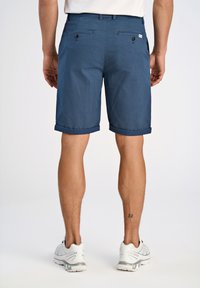 Pantaloni corti blu con un motivo testurizzato, orli risvoltati, tasche posteriori con bottoni, indossati con scarpe sportive bianche.