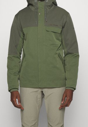 Homme portant une veste à capuche bicolore verte avec poche à rabat avant et pantalon beige sur fond uni.