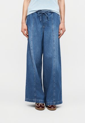 Persona con pantalones anchos de mezclilla azul con cintura de cordón y zapatos marrones, de pie contra un fondo claro y sencillo.