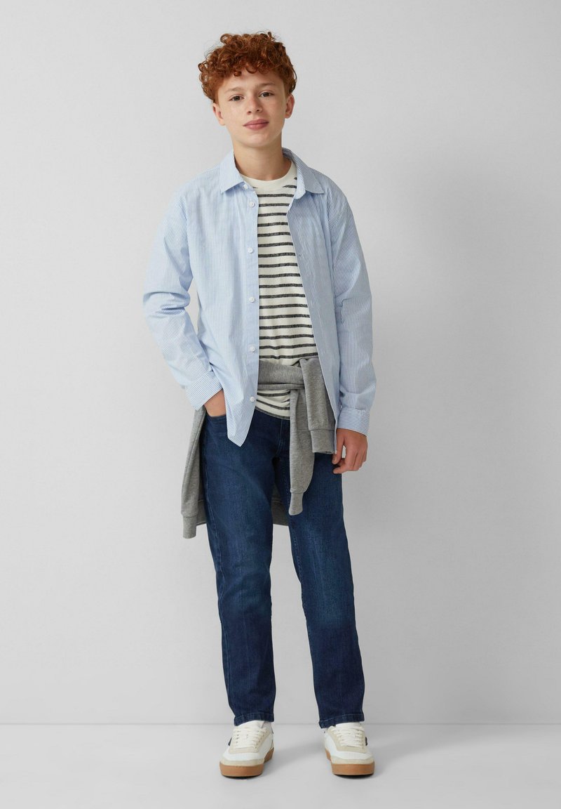 Camisa de botones a rayas azul y blanca sobre una camiseta de rayas gris, combinada con jeans oscuros y zapatillas blancas con suela marrón.