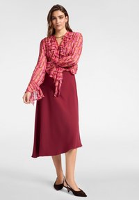 Vrouw in een gemustreerde roze blouse met ruches, een effen bordeauxrode midi-rok, zwarte puntige slingback hakken en een gouden ketting.