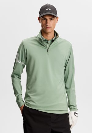 Mann trägt schwarze Mütze, hellgrünes Langarm-Oberteil mit Reißverschluss bis zur Brust, schwarze Hose und weißen Golfhandschuh, steht mit einer Hand in der Tasche.