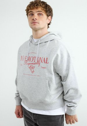 Jeune homme portant un sweat à capuche gris clair avec texte rouge "NO EMOTIONAL Responsibility" et un motif de cupidon, debout devant un fond blanc uni.