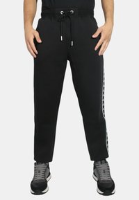 Pantaloni della tuta neri con vita elasticizzata e cordino, caratterizzati da strisce laterali con un motivo a logo bianco. Il materiale sembra morbido.