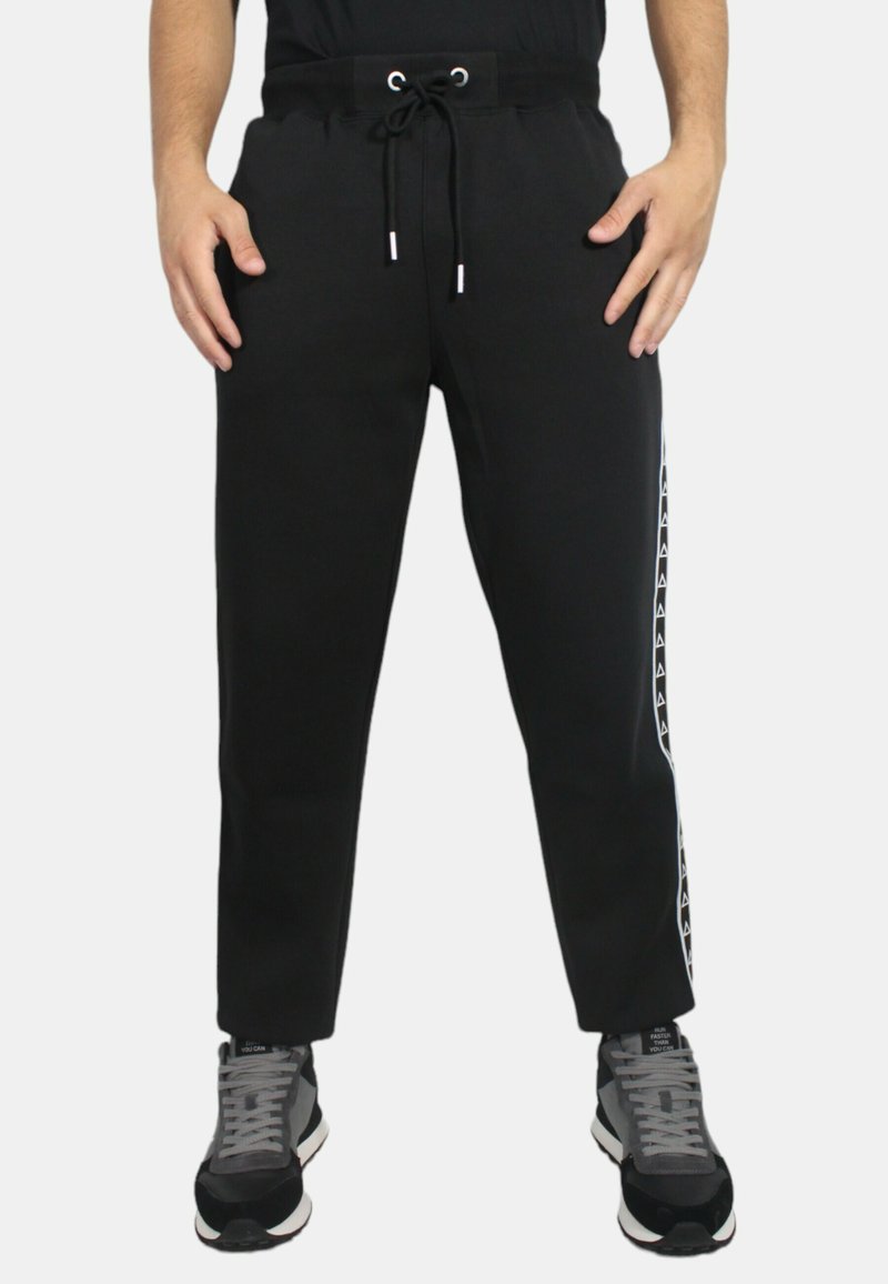 Pantaloni della tuta neri con vita elasticizzata e cordino, caratterizzati da strisce laterali con un motivo a logo bianco. Il materiale sembra morbido.