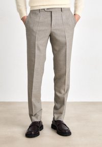 Beige kalhoty na míru s texturovanou tkaninou, s plisovaným předním dílem, bočními kapsami a rovnýma nohama, doplněné tmavými botami.