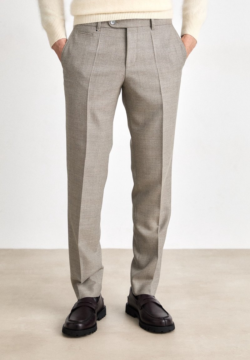 Beige kalhoty na míru s texturovanou tkaninou, s plisovaným předním dílem, bočními kapsami a rovnýma nohama, doplněné tmavými botami.