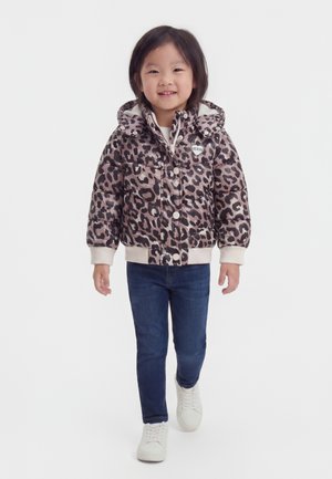 Bambino piccolo che cammina, indossa una giacca imbottita con stampa leopardata, jeans blu e sneakers bianche, sorridente su sfondo bianco semplice.