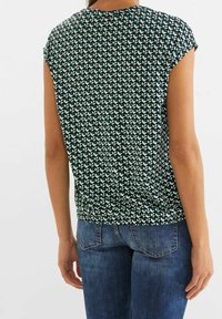 Zelený krátký rukáv s geometrickým vzorem, kulatým výstřihem a nařaseným spodním lemováním, doplněný modrými džíny.