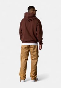 Brauner Kapuzenpullover mit großer Kapuze auf der Rückseite, kombiniert mit beigen Hosen. Das Outfit hat eine lockere Passform und sichtbare Nähte.