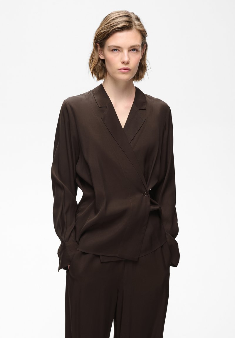 Femme aux cheveux courts portant un blazer portefeuille à manches longues marron foncé et un pantalon assorti sur un fond clair uni.