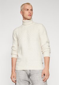 Maglione lavorato a maglia di colore crema con colletto alto a coste, motivo a spina di pesce testurizzato e polsini e orlo a coste, abbinato a jeans grigi chiari.