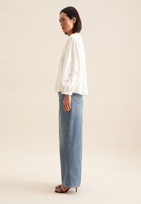 Blusa bianca a maniche lunghe con maniche ricamate, abbinata a jeans azzurri chiari a vita alta e tacchi neri.