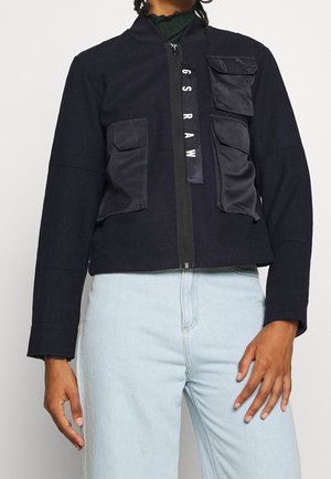 Personne portant une veste noire avec plusieurs poches avant et une fermeture éclair verticale étiquetée "G S R A W,", associée à un jean bleu clair.