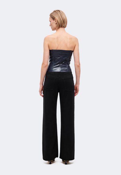 Modèle vu de dos, portant un haut noir sans bretelles à la texture travaillée, un pantalon noir à jambes larges et des chaussures à talons, sur un fond clair uni.