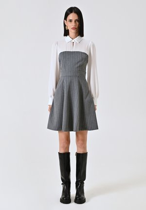 Graues Nadelstreifenkleid mit eng anliegendem Oberteil, schwingendem Rock und weißer Bluse mit hohem Kragen. Kombiniert mit hohen schwarzen Stiefeln.