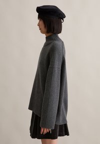 Γυναίκα σε προφίλ που φοράει ένα χαλαρό σκούρο γκρι πουλόβερ με ψηλό γιακά, μαύρη μπερέ και μαύρη φούστα, απέναντι από απλό φόντο.