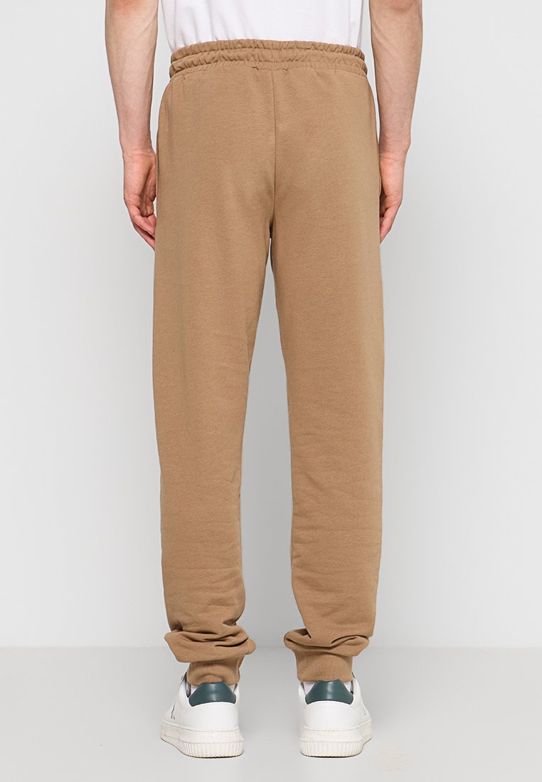 Pantaloni della tuta marroni con una texture morbida, cintura elastica e polsini affusolati; indossati con scarpe da ginnastica bianche con un accento verde.