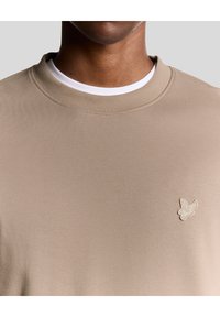 Beige sweatshirt med rund hals lavet af glat stof, med et lille broderet logo på venstre bryst og en lagdelt hvid krave.