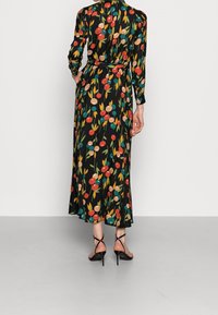 Žena v dlouhých černých květovaných šatech se červenými, zelenými a béžovými kruhovými vzory, doplněná černými sandály na vysokém podpatku, stojící s jednou rukou v kapse.