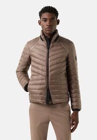 Beige donsjas met doorgestikte structuur, hoge kraag, zwarte ritssluiting en zijzakken. Beschikt over een logo op de linker mouw als detail.