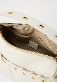 Borsa in finta pelle color crema con accessori in oro, chiusura con zip e interno foderato in satin. Presenta una tracolla robusta con occhielli decorativi.