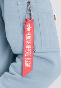 Plava dukserica s metalnim zatvaračem i crvenom oznakom "UKLONI PRIJE LETA". Glatko platno i istaknuti detalji šivanja.