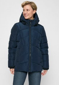 Lächelnde Frau in einer navy-blauen, gefütterten Jacke mit hohem Kragen und Kapuze, kombiniert mit blauen Jeans, vor einem weißen Hintergrund.