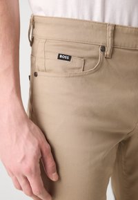 Beige broek met een slanke pasvorm, voorzien van een klein zwart "BOSS"-label, voorzakken en subtiele stiksel-details. Glad stoftextuur.