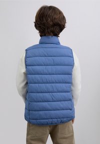 Chaleco acolchado azul con costuras horizontales, cuello alto y logo en el cuello. Llevado sobre una camisa blanca de manga larga, combinado con pantalones beige.