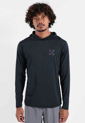 Homme aux cheveux bouclés portant un sweat à capuche noir à manches longues avec un petit logo géométrique violet sur la poitrine, debout devant un fond uni.