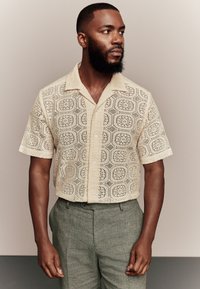 Koronkowa koszula z krótkim rękawem w jasnobeżowym kolorze z geometrycznymi wycięciami, zestawiona z szarymi lnianymi spodniami. Guziki z przodu.