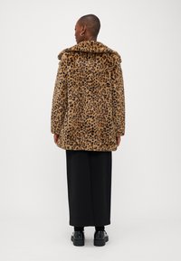 Kaput s imitacijom leopard uzorka od lažnog krzna, sa širokim kragnom, ravnim krojem i prednjim kopčanjem na dugmad. Crne hlače širokog kroja i robusne cipele zaokružuju izgled.