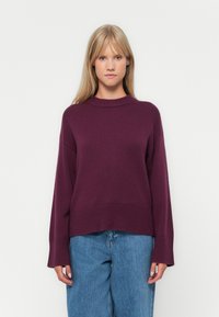 Maglione viola lavorato a maglia con colletto alto, spalle scese e orlo a coste. Indossato con jeans in denim blu larghi.