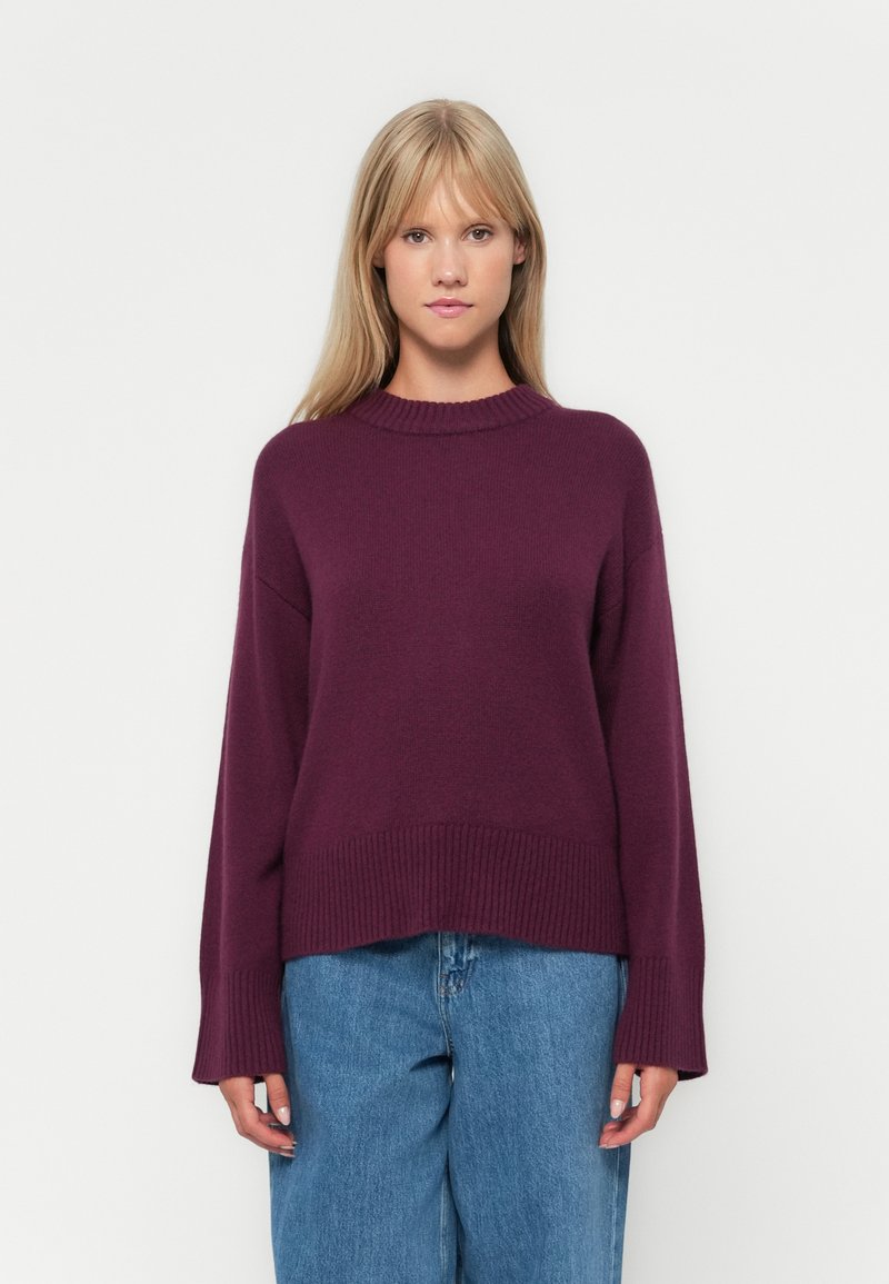 Lila gestrickter Pullover mit hohem Kragen, überschnittenen Schultern und geripptem Saum. Getragen mit lockeren blauen Jeans.
