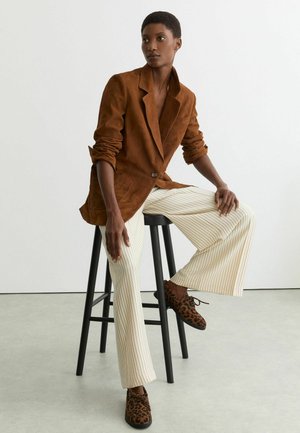 Frau sitzt auf einem schwarzen Hocker und trägt ein braunes Blazer, weiße, breitbeinige Hosen mit Nadelstreifen und Schuhe mit Leopardenmuster vor weißem Hintergrund.