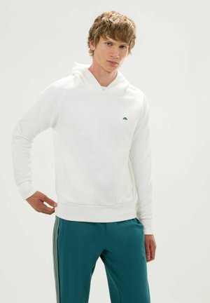 Joven con cabello castaño claro, vestido con una sudadera blanca con capucha y pantalones azul verdoso con rayas blancas, de pie frente a un fondo claro y liso.