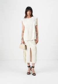 White, fringed two-piece outfit with asymmetrical design. Features a sleeveless top and pencil skirt with slit. Black high-heeled sandals and clutch.