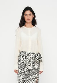 Μπεζ μακρυμάνικο πουκάμισο με στρογγυλό λαιμόκοψη και κούμπωμα με κουμπιά, συνδυασμένο με φούστα τύπου περιτύλιγμα σε ασπρόμαυρο μοτίβο με φιόγκο.