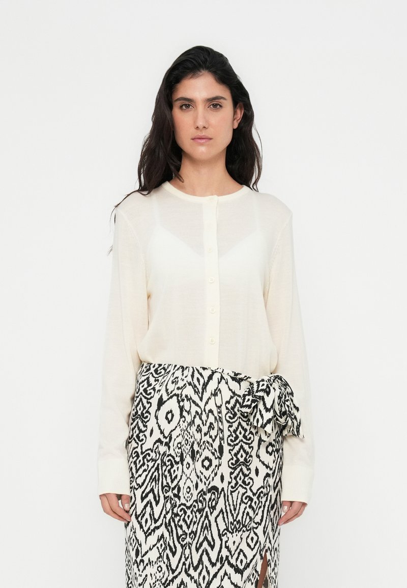 Μπεζ μακρυμάνικο πουκάμισο με στρογγυλό λαιμόκοψη και κούμπωμα με κουμπιά, συνδυασμένο με φούστα τύπου περιτύλιγμα σε ασπρόμαυρο μοτίβο με φιόγκο.