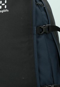 Svart och mörkblå ryggsäck med en texturerad yta, justerbara remmar och en plastspänne. Logotypen syns på framsidan.