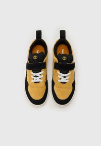 Baskets en daim jaune avec accents noirs, lacets blancs et sangle velcro. Design léger avec des matériaux texturés et un col rembourré.