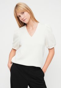 Jeune femme aux cheveux blonds lisses portant un chemisier blanc à manches bouffantes et encolure en V, et un pantalon noir, debout les mains dans les poches.